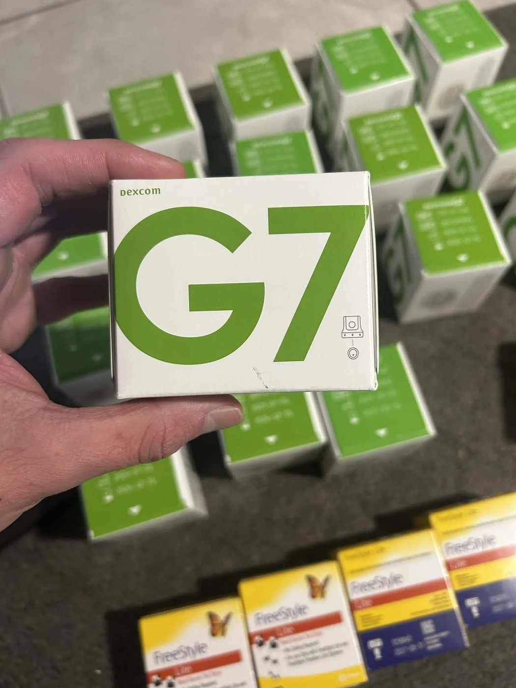 A sealed Dexcom G7 sensor box held in front of dozens more G7 and FreeStyle Lite boxes